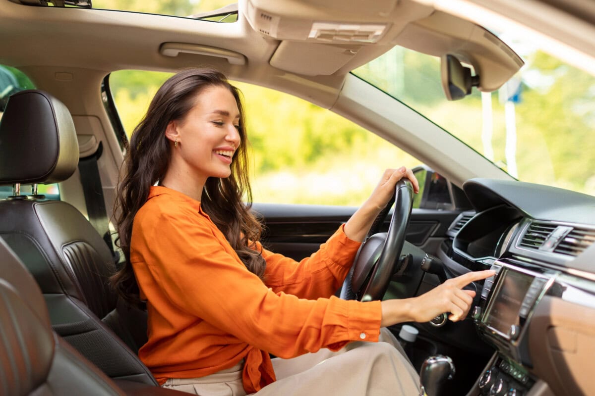 Happy woman driving car and listening to music radio on auto audio system, checking auto options during test drive, pushing button on dashboard